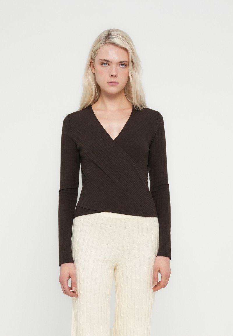 Maglia marrone a maniche lunghe con costine, scollo a V e design avvolgente, abbinata a pantaloni a gamba larga color crema con lavorazione a trecce.