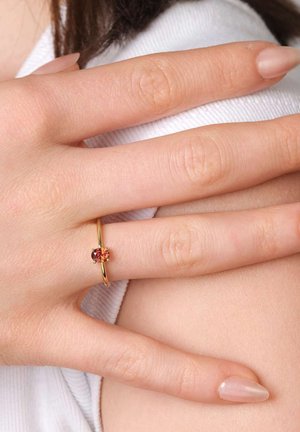 Hand with polished nails wearing a thin gold ring featuring a small red gemstone, resting on a white fabric background.