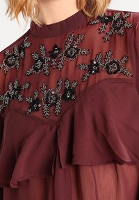 Blouse couleur vin featuring tissu transparent, broderie florale noire détaillée, une couche à volants et un col montant.