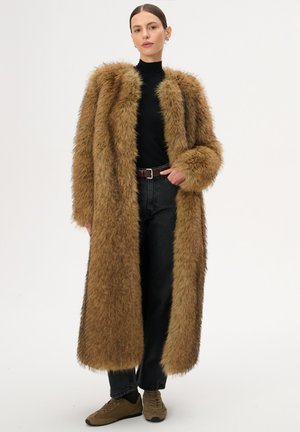 Kobieta ubrana w długi brązowy płaszcz z futra ekologicznego, czarny golf, czarne spodnie, brązowy pasek i brązowe trampki, stojąca z jedną ręką w kieszeni.