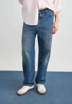 Άτομο που φοράει ξεθωριασμένα μπλε τζιν παντελόνια ευθείας γραμμής, λευκά αθλητικά παπούτσια με μπεζ αποχρώσεις, και ένα ανοιχτό ροζ πουκάμισο που στέκεται σε μπλε χαλί.