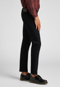 Černé džíny s úzkým střihem, s texturovaným povrchem a logem na pasu. Spárovány s černými šněrovacími botami.