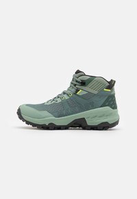 Scarpa da hiking verde con tomaia in mesh testurizzato, suola in gomma nera, sistema di allacciatura distintivo e logo "MAMMUT" sul lato. Presenti dettagli gialli.