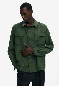 Green camo-patterned button-up shirt with two chest pockets, a classic collar, and a relaxed fit. Features a smooth texture and shiny buttons.