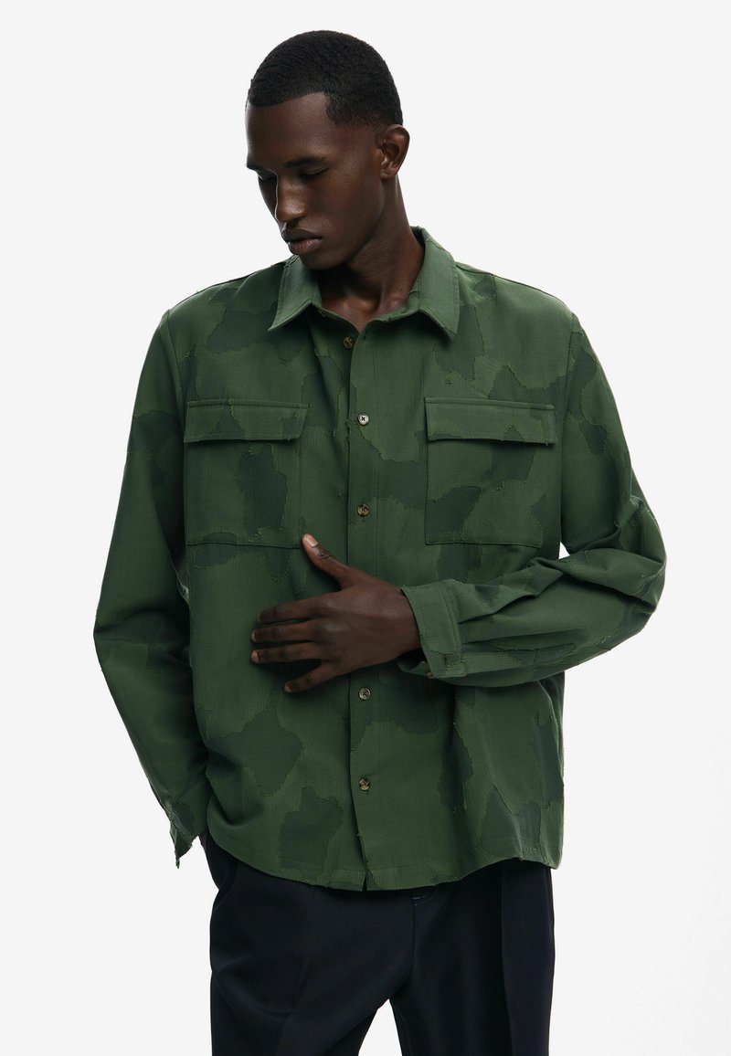 Green camo-patterned button-up shirt with two chest pockets, a classic collar, and a relaxed fit. Features a smooth texture and shiny buttons.