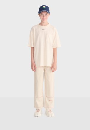 Giovane che indossa una T-shirt oversize color crema e pantaloni a gamba larga abbinati con cuciture rosse, scarpe bianche divise alle dita e un berretto blu con grafica floreale.