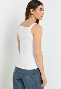 Camisola bianca a coste con scollatura quadrata e spalline sottili. Il tessuto sembra morbido ed elasticizzato. Indossata con jeans a vita alta.