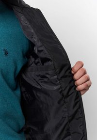 Hånd som holder åpen svart jakke med innvendig lomme, blågrønn genser med lite brodert logo synlig under.