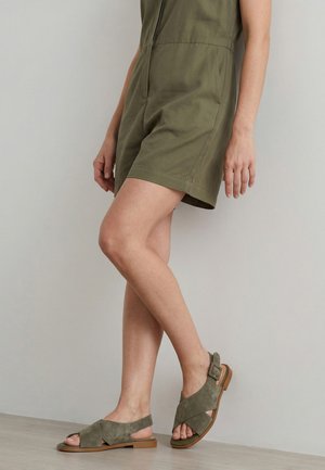 Persoon die een olijfgroene romper en bijpassende open sandalen met enkelriem draagt, staat met gekruiste benen tegen een neutrale muur.