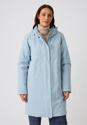Femme portant un manteau d'hiver bleu clair à capuche par-dessus un col roulé beige et un pantalon à carreaux foncé, debout devant un fond gris uni.
