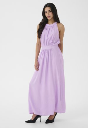 Mujer con un vestido maxi lavanda sin mangas y cintura fruncida, de pie y posando con tacones negros puntiagudos sobre un fondo blanco.