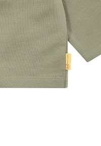 Maglietta a maniche lunghe verde a coste con orlo dritto, che presenta una piccola etichetta gialla sul lato con dettaglio del logo circolare.