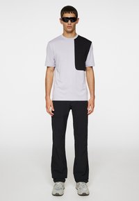 Ljusgrå t-shirt med svart accent på axeln, kombinerad med svarta byxor. Modellen bär mörka solglasögon och grå sneakers.