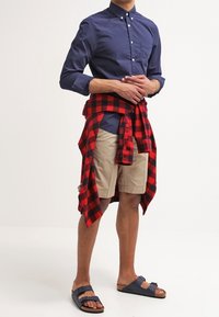 Un homme portant une chemise boutonnée bleu marine, un short beige, des sandales bleu marine et une chemise à carreaux rouge et noir nouée autour de la taille.