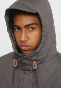 Kaptur z wysokim kołnierzem wykonany z stonowanej szarej tkaniny, z ozdobnymi elementami i sznurkiem przy twarzy.