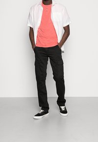 Černé cargo kalhoty, korálové henley tričko a přes ně bílá krátký rukáv. Černé tenisky s bílými podrážkami. Jednoduchý design.