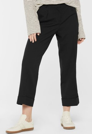 Person trägt schwarze, gekürzte Hosen mit Umschlägen, ein langärmliges beige-schwarz gestreiftes Oberteil und weiße Sneakers mit Gummisohlen.