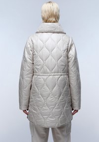 Giacca trapuntata beige chiaro con colletto alto, caratterizzata da una texture a onde e una vita sfiancata. Maniche lunghe con vestibilità rilassata.
