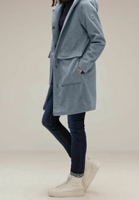 Ljusblå herringbone-kappa med huva, med två framfickor, knäppning och textilstruktur, i kombination med mörka jeans och vita sneakers.