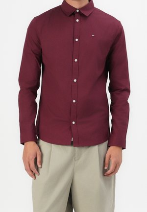 Mężczyzna ubrany w dopasowaną, bordową koszulę na guziki z długim rękawem oraz jasnobeżowe spodnie, stojący na tle jednolitego, białego tła.