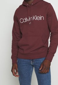 Marsaľový mikina s bielym logom "Calvin Klein". Obsahuje kapuci s šnúrkou, klokanie vrecko a žebrované manžety a lem.