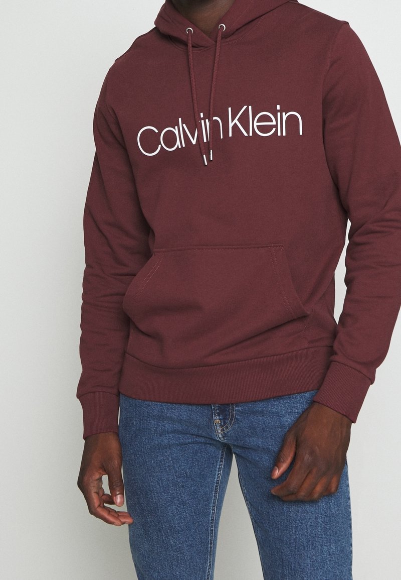 Marsaľový mikina s bielym logom "Calvin Klein". Obsahuje kapuci s šnúrkou, klokanie vrecko a žebrované manžety a lem.