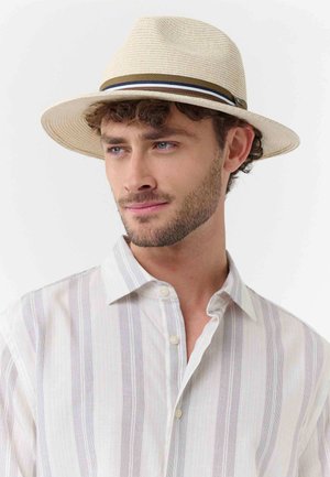 Junger Mann mit lockigem Haar, der einen beigen Strohhut mit gestreiftem Band und ein weißes Hemd mit vertikalen Streifen trägt.