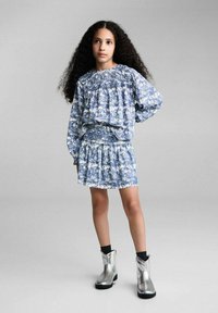 Blauwe jurk met bloemenpatroon, met pofmouwen en gathered taille, gecombineerd met glanzende zilveren enkellaarsjes en zwarte sokken.