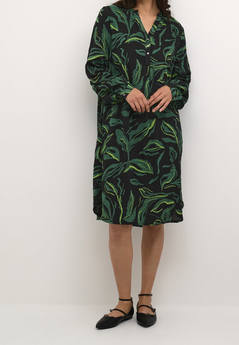 Femme portant une robe noire à manches longues avec des motifs de feuilles vertes et des chaussures plates noires à bride à la cheville, debout contre un mur blanc uni.