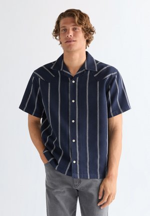 Nooremees lokkis juustega kannab tumesinist lühikeste varrukatega triiksärki valgete vertikaalsete triipudega ja halli pükse, seistes ühe käega taskus.