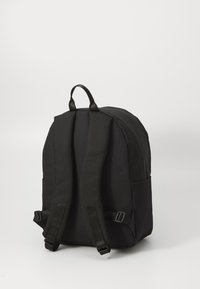 Mochila de tela negra con correas ajustables, asa en la parte superior y bolsillos laterales. Presenta una forma redondeada y una textura suave.