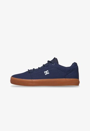 Navy canvas sneakers with a tan rubber sole, featuring a white DC logo, rounded toe, and lace-up design. Flat textile tongue and collar.