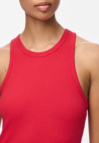 Kobieta w bezrękawnym, czerwonym, prążkowanym topie, ukazująca szyję i górną część torsu na białym tle.