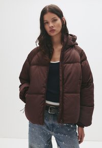 Gepolsterte, kapuzenjacke in Dunkelbraun mit gestepptem Design. Verfügt über einen Frontreißverschluss und seitliche Taschen; getragen über einem marineblauen Oberteil und zerrissenen Jeans.