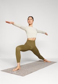Olive green high-waisted leggings and a cream ribbed long-sleeve crop top. The mat has a textured, speckled surface. Model in a warrior pose.