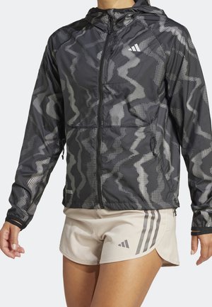 Person trägt eine schwarze, gemusterte Kapuzenjacke mit Reißverschluss und hellbeige Shorts mit grauen Adidas-Logos und -Streifen, steht mit entspannten Armen.