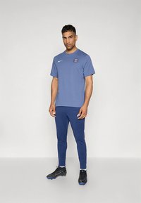 Nike športna obleka, ki vključuje modro majico s kratkimi rokavi in logotipom ter ujemajoče se modre oprijete hlače, skupaj s črnimi športnimi čevlji.