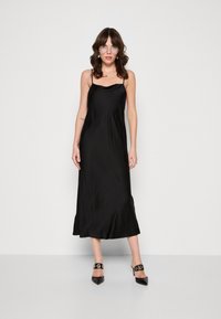 Robe nuisette en satin noir avec des bretelles délicates, présentant un drapé doux et un ourlet modeste. Assortie à des talons aiguilles noirs.