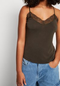 Olivgrön linne med spetsdetalj vid halsringningen, med tunna axelband och en slät, figurnära design. Matchas med ljusblå jeans.