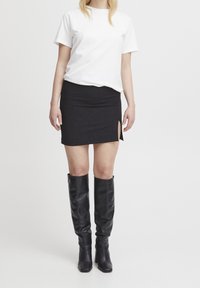 Žena v jednoduchém bílém tričku s krátkým rukávem, černé mini sukni s bočním rozparkem a černých kozačkách na vysokém podpatku stojí na bílém pozadí.