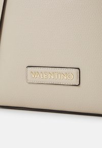 Beige läderväska med en texturerad yta, svart kantdetalj och en guldfärgad "Valentino"-logotyp på en rektangulär tag.