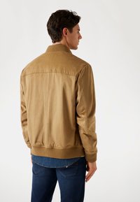 Tan bomberjack met een gladde textuur en geribde kraag, weergegeven vanaf de achterkant, heeft een aansluitende tailleband en een minimalistisch ontwerp.