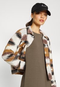 Jaqueta de flanela com padrão geométrico em castanho, branco e preto; design cropped; fecho com botões; combinada com uma blusa cinza de mangas longas e um boné preto.