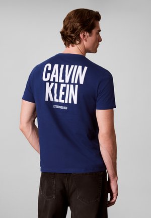 Mand set fra siden iført en blå t-shirt med stor hvid "Calvin Klein" tekst og "Established 1968" på ryggen, kombineret med sorte bukser.