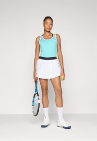 Tennisoutfit mit einem mintgrünen Tanktop mit weißem Saum, einem plissierten weißen Rock und weißen Schuhen. Das Model hält einen Tennisschläger und einen Ball.