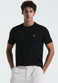 Zwart T-shirt met korte mouwen, gemaakt van zacht fabric, met een ronde halslijn en een klein wit logo op de linkerborst. G gedragen met lichte broek.