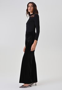 Top nero a maniche lunghe con dettagli trasparenti abbinato a pantaloni larghi in velluto nero a vita alta; modella indossa scarpe con tacco metallico.