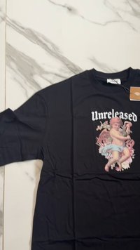 Černé tričko s centrálním grafickým motivem cherubínské postavy obklopené květinami, s nápisem "Unreleased" nahoře v bílé barvě.
