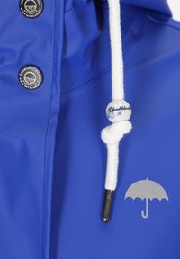Kék vízálló dzseki, textúrált felülettel, fehér húzózsinórral és gyöngyökkel, valamint kör alakú fekete gombokkal. Tartalmaz egy esernyő logót.