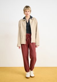 Beige leichte Jacke mit einem Kragen, kombiniert mit bordeauxfarbenen strukturierten Jogginghosen und weißen Turnschuhen mit beigen Sohlen.